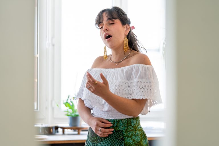 A young woman passionately singing indoors, wearing a white top with green skirt.